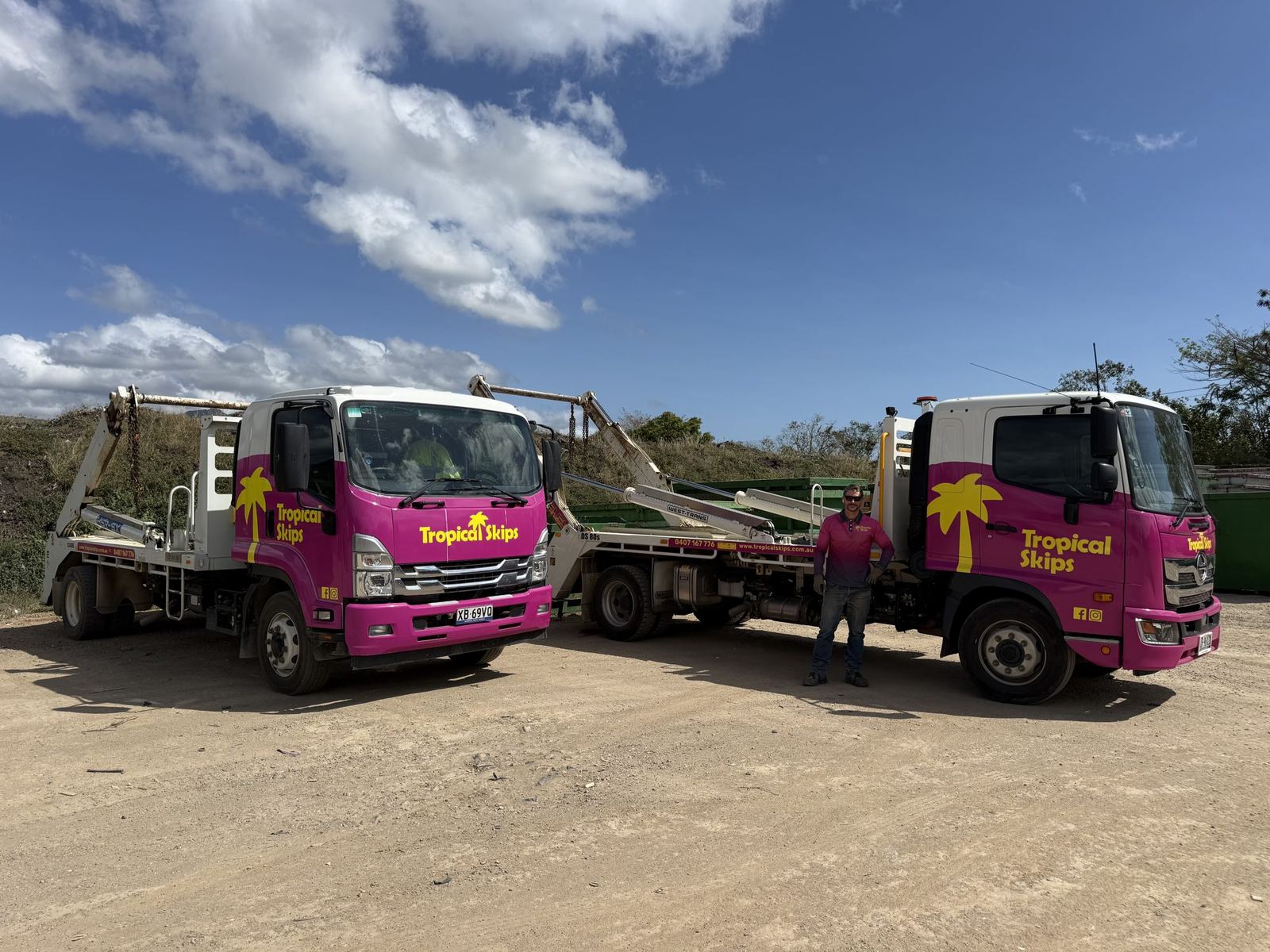 Two Tropical Skips trucks parked side by side at the yard, hills behind